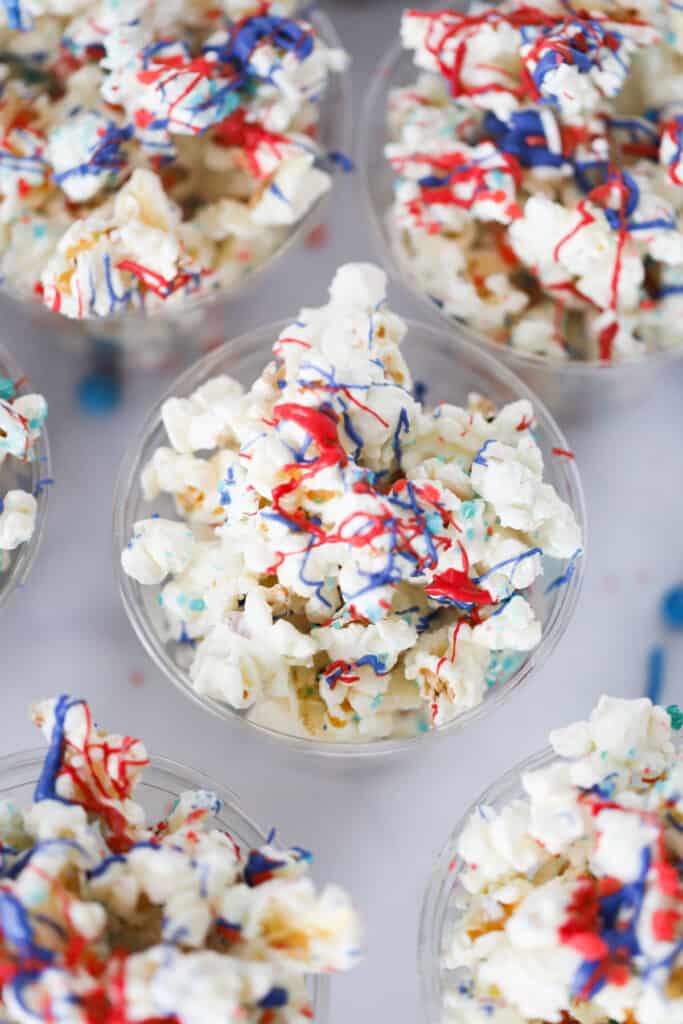 Firecracker Popcorn inside single serving plastic cups, topped with blue and red drizzle. sprinkles popcorn.
