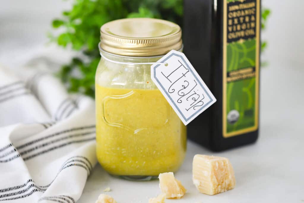 A mason jar full of dressing with a tag that says "Italian" on a table next to a jar of olive oil, a fresh chunk of Parmesan cheese and a towel; making Italian Dressing.