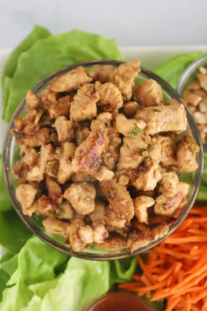 A small bowl full of cooked chicken, surrounded by lettuce leaves, sauces, and shredded carrots to make lettuce wraps.