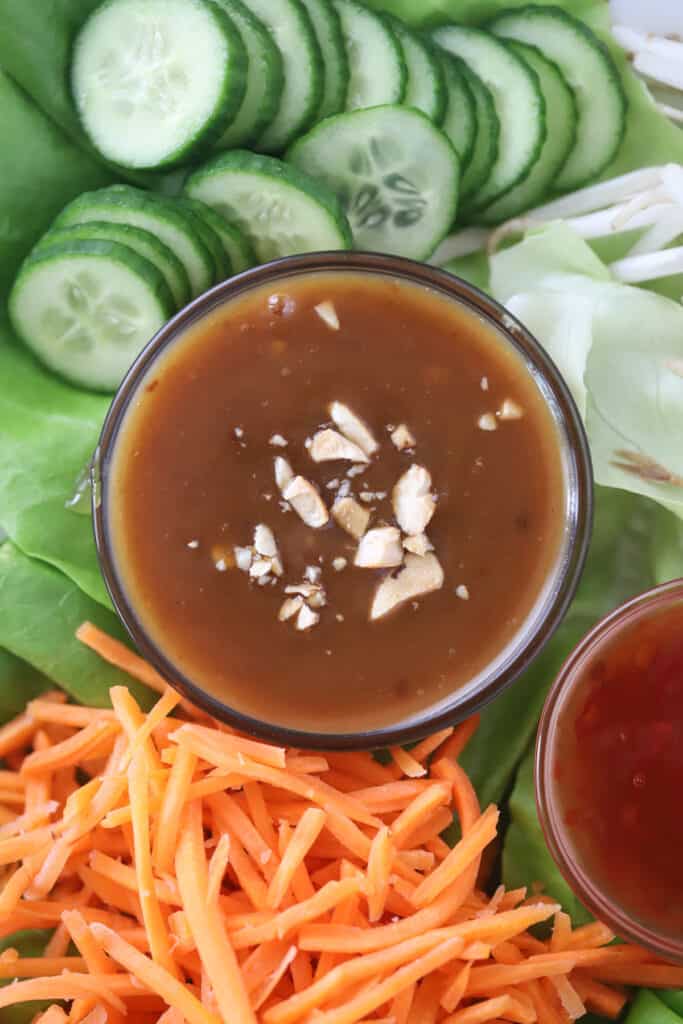 A ramekin of peanut sauce chicken lettuce wraps on a platter with sliced cucumbers, lettuce leaves, and shredded carrots.