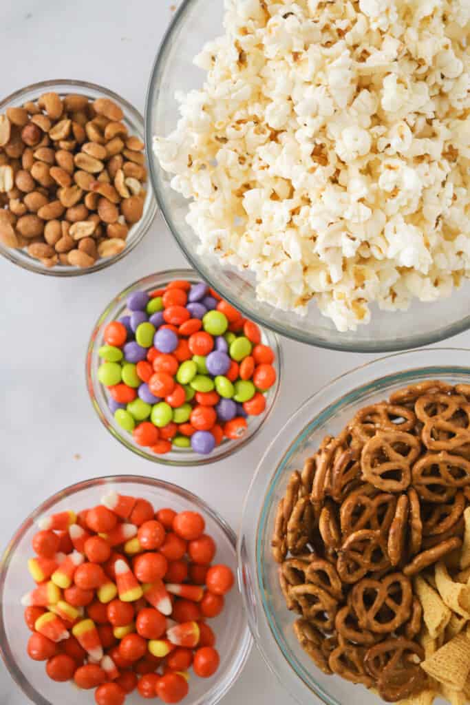 Small bowls full of ingredients to make this party mix recipe, including peanuts, popcorn, M&Ms, pretzels, and candy corn.
