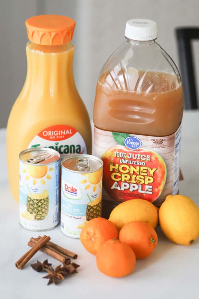 Ingredients to make this Wassail recipe on a table, including orange juice, apple juice, pineapple juice, spices, and whole citrus. wassail recipe alcohol, traditional wassail recipe 