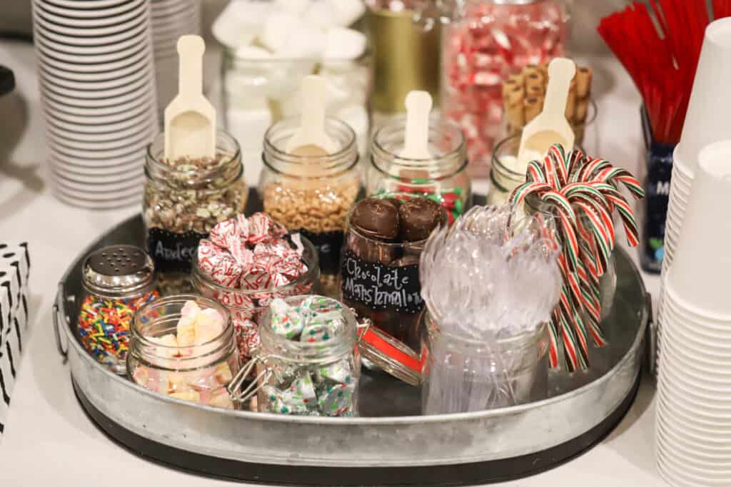 A hot chocolate bar set up on a table, complete with cups and a serving platter full of mason jars with toppings and scoops. ideas for hot chocolate bar. 