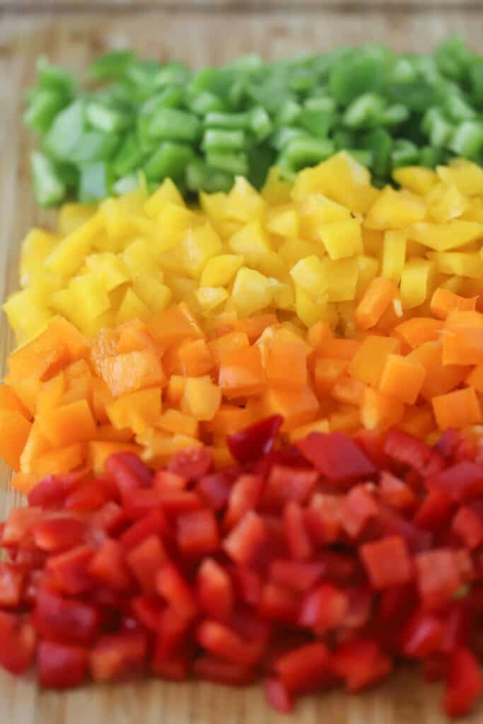 Green, yellow, orange, and red bell peppers diced and laying on a cutting board in rows.