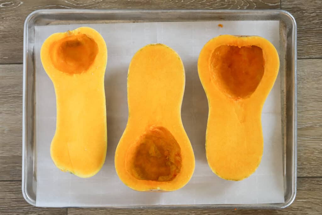 A sheet pan lined with parchment paper that has three halves of butternut squash with the seeds scooped out. Butternut squash cooking time, baking butternut squash halves.