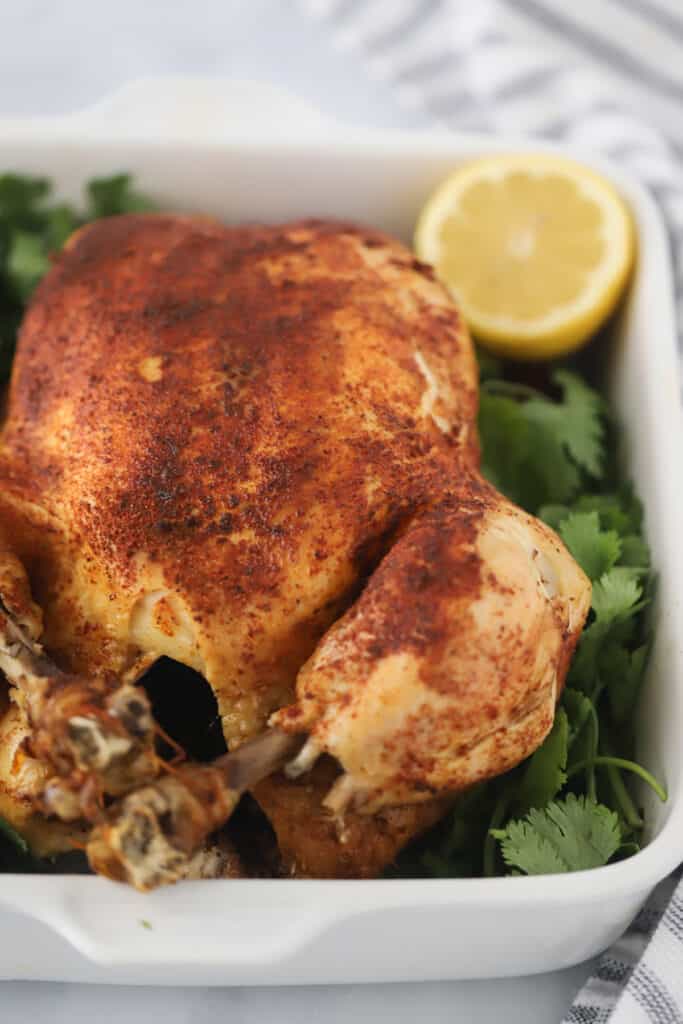 A whole cooked chicken in a baking dish surrounded by fresh parsley and lemon wedges.
