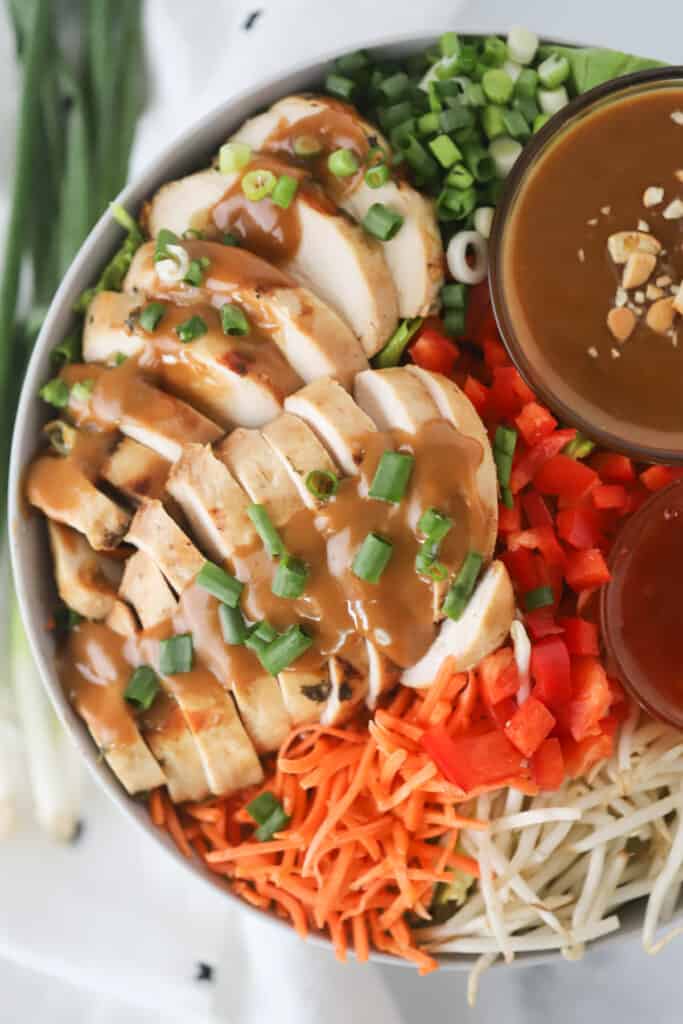 A large bowl full of dipping sauces, shredded carrots, bean sprouts, sliced green onions, and sliced Thai chicken covered in peanut sauce.