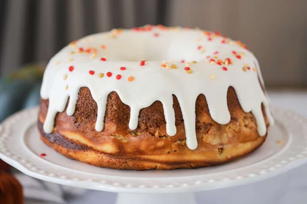 A Pumpkin Bundt Cake topped with glaze and sprinkles on a white serving plate. Pumpkin cake ideas, easy pumpkin cake recipe.