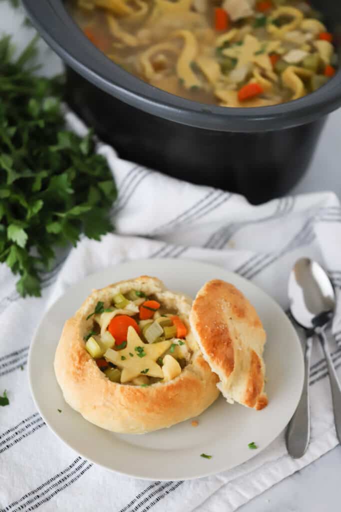 A white serving plate with a bread bowl full of chicken noodle soup.