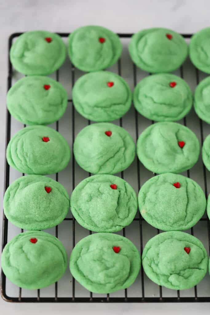 A wire rack full of green Grinch Sugar Cookies with a small candy heart stuck in each.