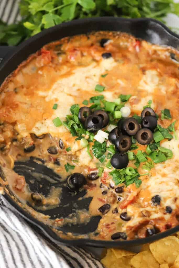 A cast iron skillet full of Cowboy Queso topped with olives, cilantro, and green onions. A baked queso dip with ground beef.