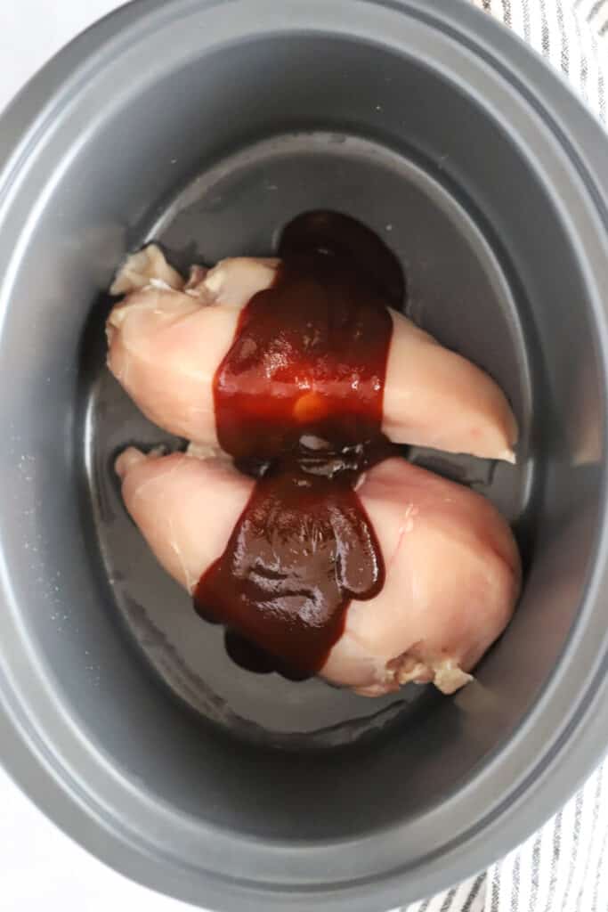 A crockpot with raw chicken breast covered in BBQ sauce. slow cooker bbq chicken sandwiches. 