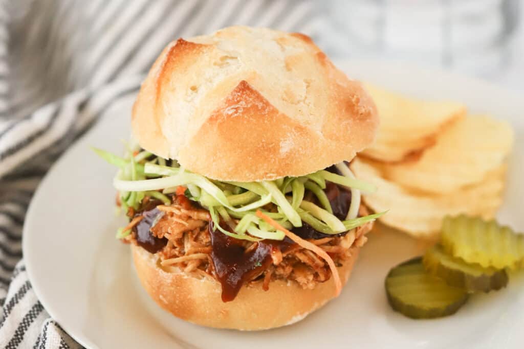 A plate with a pull chicken sandwich, chips, and pickles. how to make slow cooker bbq chicken sandwiches. Slow Cooker bbq chicken breast. Slow cooker bbq chicken recipes.