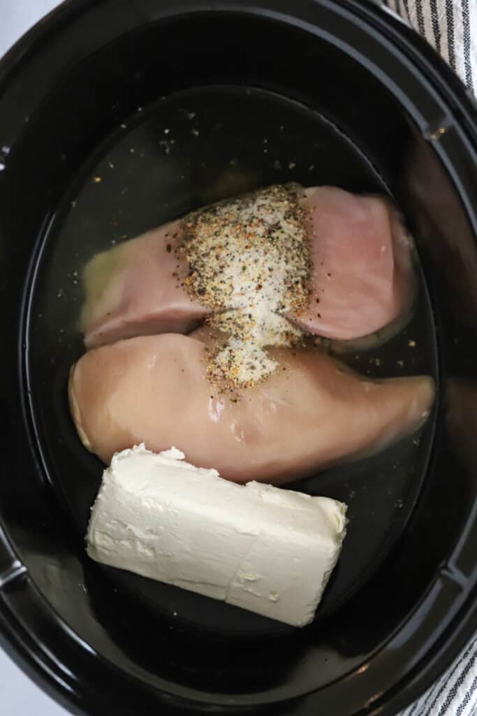 A slow cooker with raw chicken breasts, cream cheese, and seasoning ready to cook. 