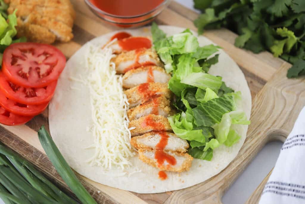 A tortilla laying flat with chicken, cheese, and lettuce ready to be turned into a buffalo chicken wrap. Buffalo chicken wraps recipe.