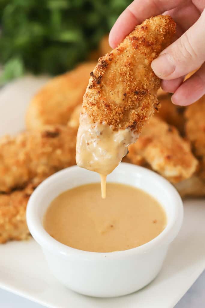 A hand holding an Air Fryer Chicken Tender and dipping it into a cup of sauce.