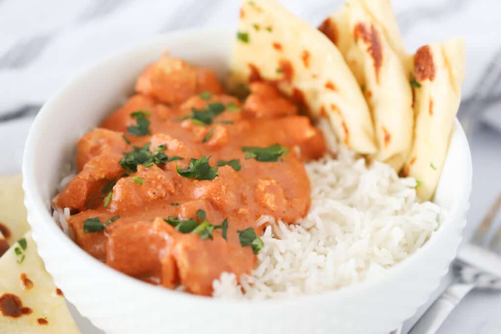 A white serving bowl full of Chicken Tikka Masala, rice, and Naan.