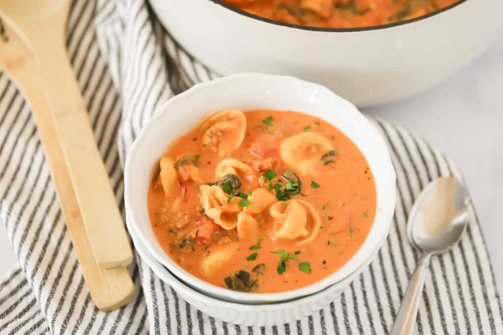 A bowl of Creamy Sausage Tortellini Soup on a table with a spoon and decorative cloth napkin. tortellini soup creamy. Tortellini soup recipes. Creamy sausage soup.