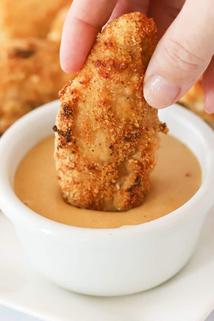 A hand dipping a chicken tender into a ramekin of sauce.