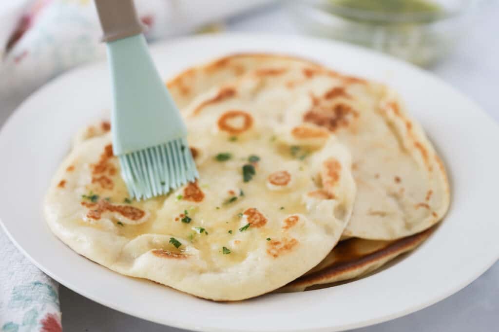 A white plate full of baked naan with melted butter being brushed on the top. Best homemade naan bread recipe.