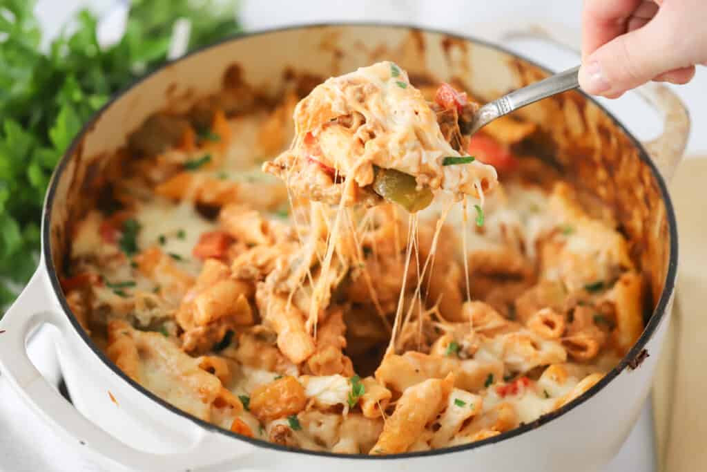 A handle scooping out a portion of a baked pasta full of cheesy sausage and peppers pasta. Cheesy sausage pasta bake.