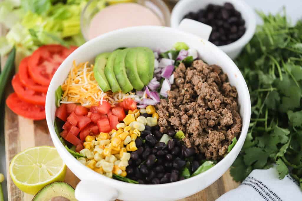 A bowl full of ingredients to make a salad including ground beef, black beans, corn, tomatoes, shredded cheese, and avocados. Easy Taco Salad Recipe. Beef taco salad.