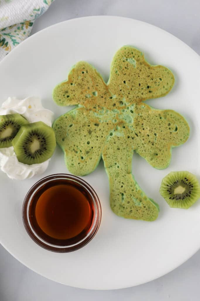 A white plate with a green shamrock shaped pancake and a side of syrup and sliced kiwi.