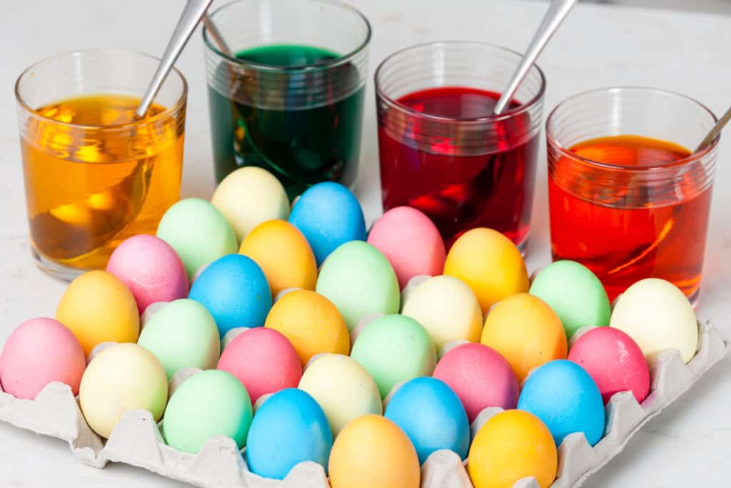 A full carton of colorful Easter Eggs with cups of dye behind; dyed Easter eggs; how to dye Easter eggs with food coloring.