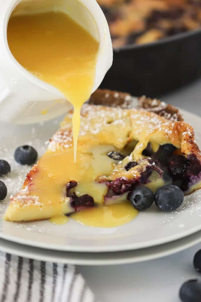 A slice of a Lemon Blueberry German Pancake on a plate with lemon sauce poured over the top; best blueberry dutch baby recipe. Lemon blueberry dutch babies.