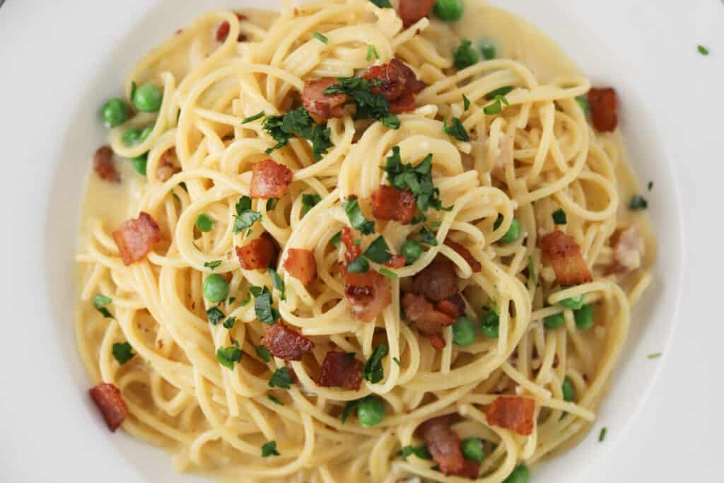 A bowl full of Spaghetti alla Carbonara topped with parsley, bacon, and peas.