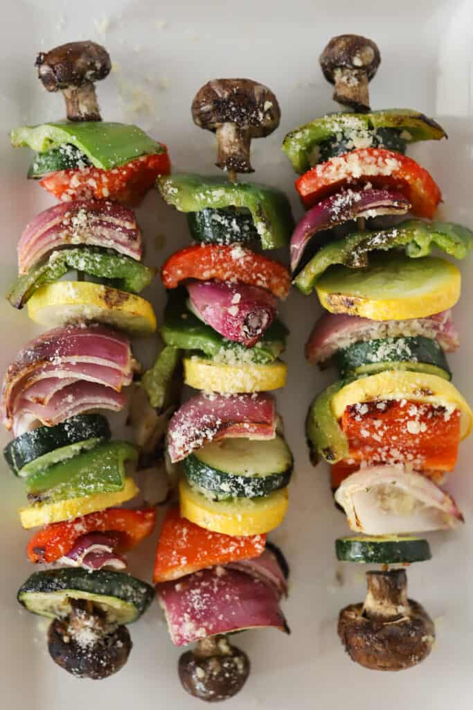 Three veggie kabobs lined up on a serving plate, sprinkled with parmesan cheese.