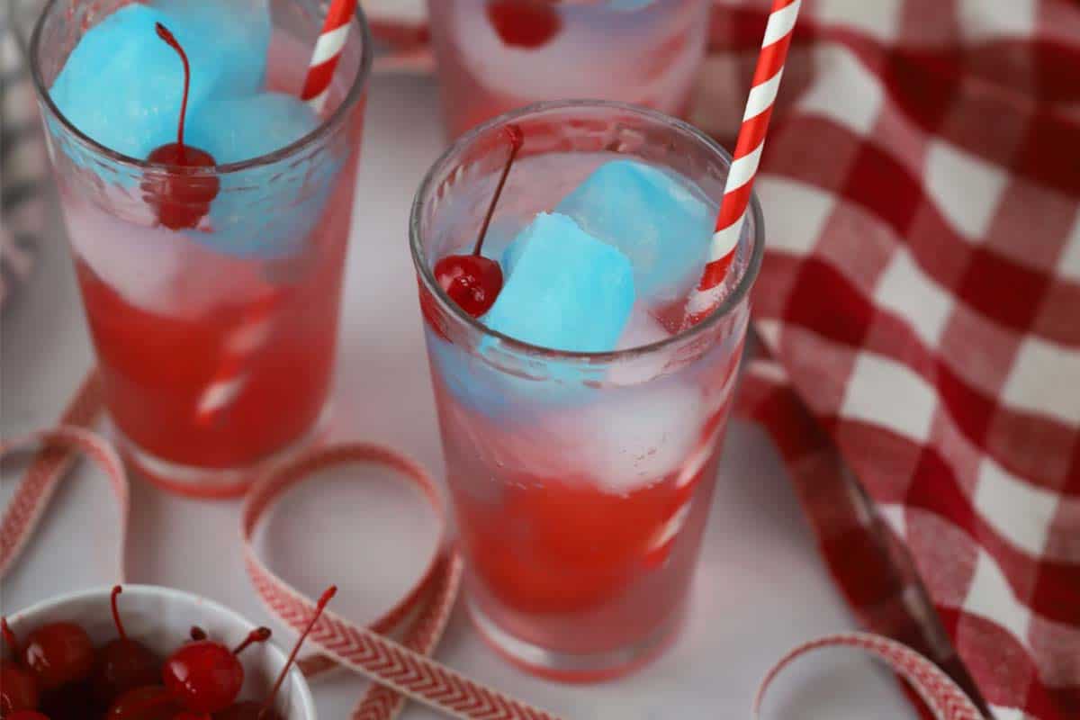 Patriotic Zeal 3 Two glasses with red white and blue 4th of july drinks made with colorful ice cubes of gatorade and maraschino cherries. The glasses are on a wooden table with a blurred festive background.