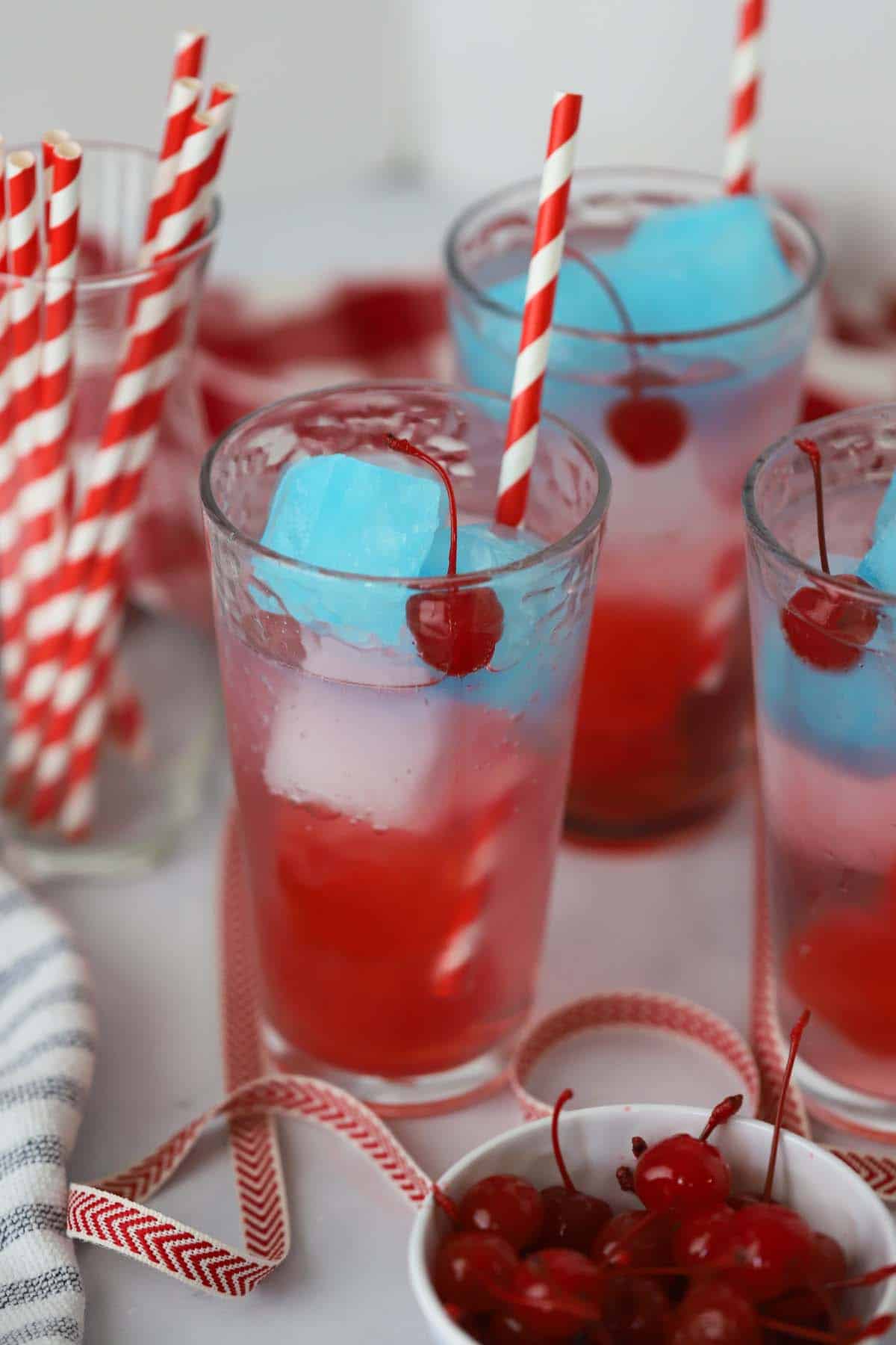 Patriotic Zeal 2 Patriotic punch, best fourth of july drinks, 4th of july drinks non alcholic. A glass filled with red, white, and blue layers of ice cubes and sparkling soda, garnished with a maraschino cherry.