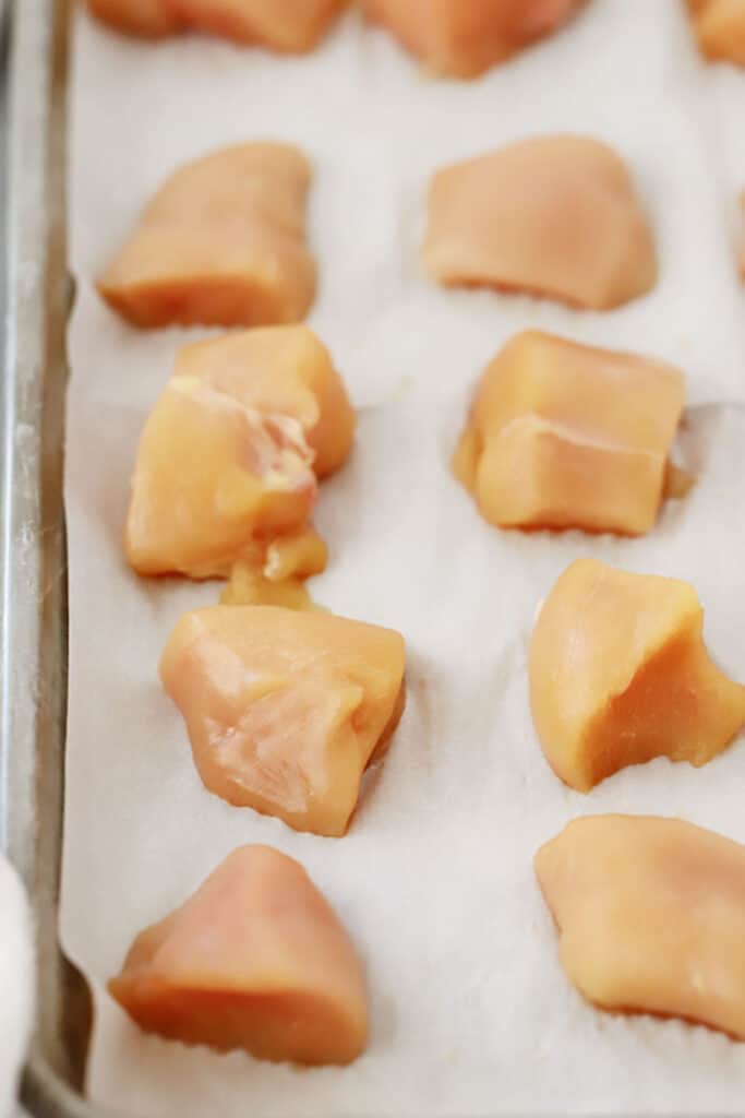 A sheet tray lined with parchment paper filled with small pieces of chicken. baked buffalo chicken breast cut into bites. Oven baked chicken bites.