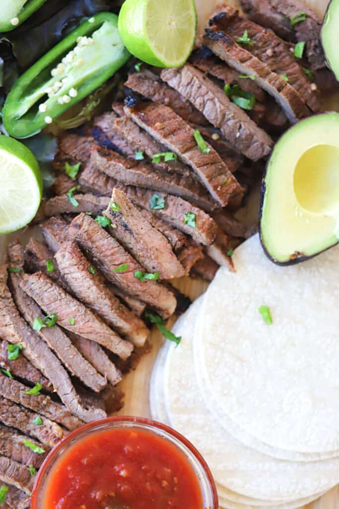 A cutting board with sliced carne asada, tortillas, and taco toppings.