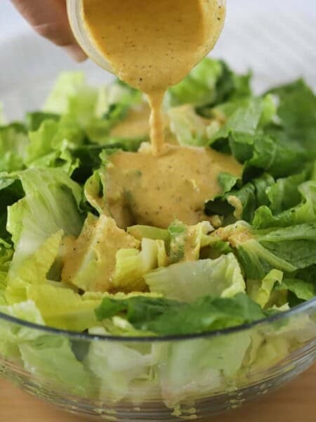 A glass bowl full of salad with homemade caesar dressing being poured over the top.