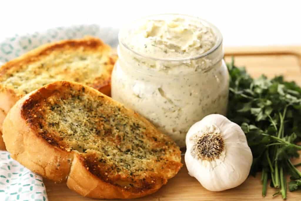 A jar full of garlic butter spread recipe next to garlic bread on a cutting board. How to make homemade garlic butter.