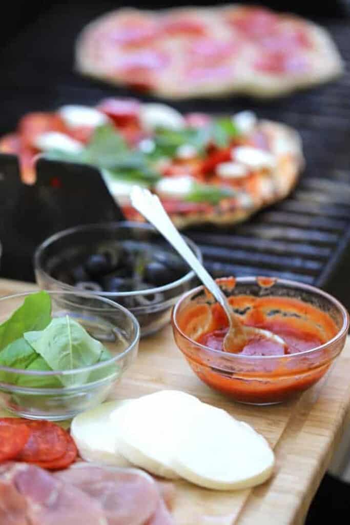 A grill cooking Grilled Pizza dough and a cutting board to the side with pizza toppings. 