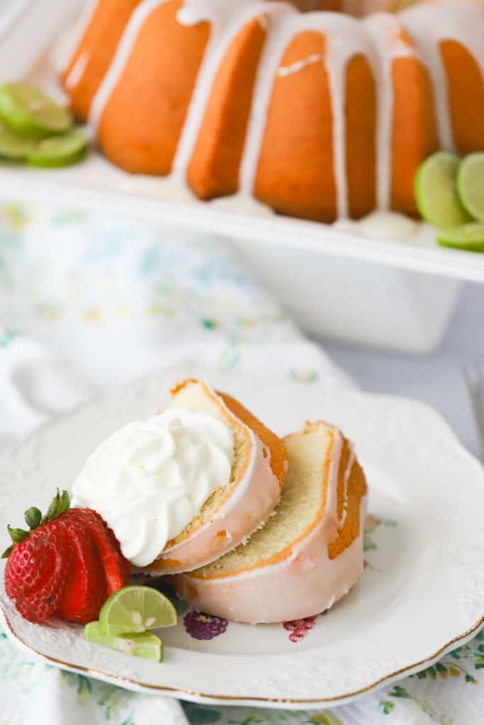 A serving plate with glazed Key Lime Pound Cake, topped with whipped cream and fresh strawberries.  A full pound cake is in the background on a cake stand. recipe for key lime pound cake. 