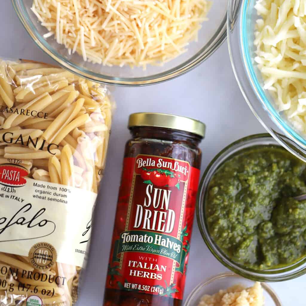 A bag of dried pasta, a jar of sun-dried tomatoes and a glass bowl full of shredded cheese on a tabletop. Chicken sun dried tomato pasta bake.