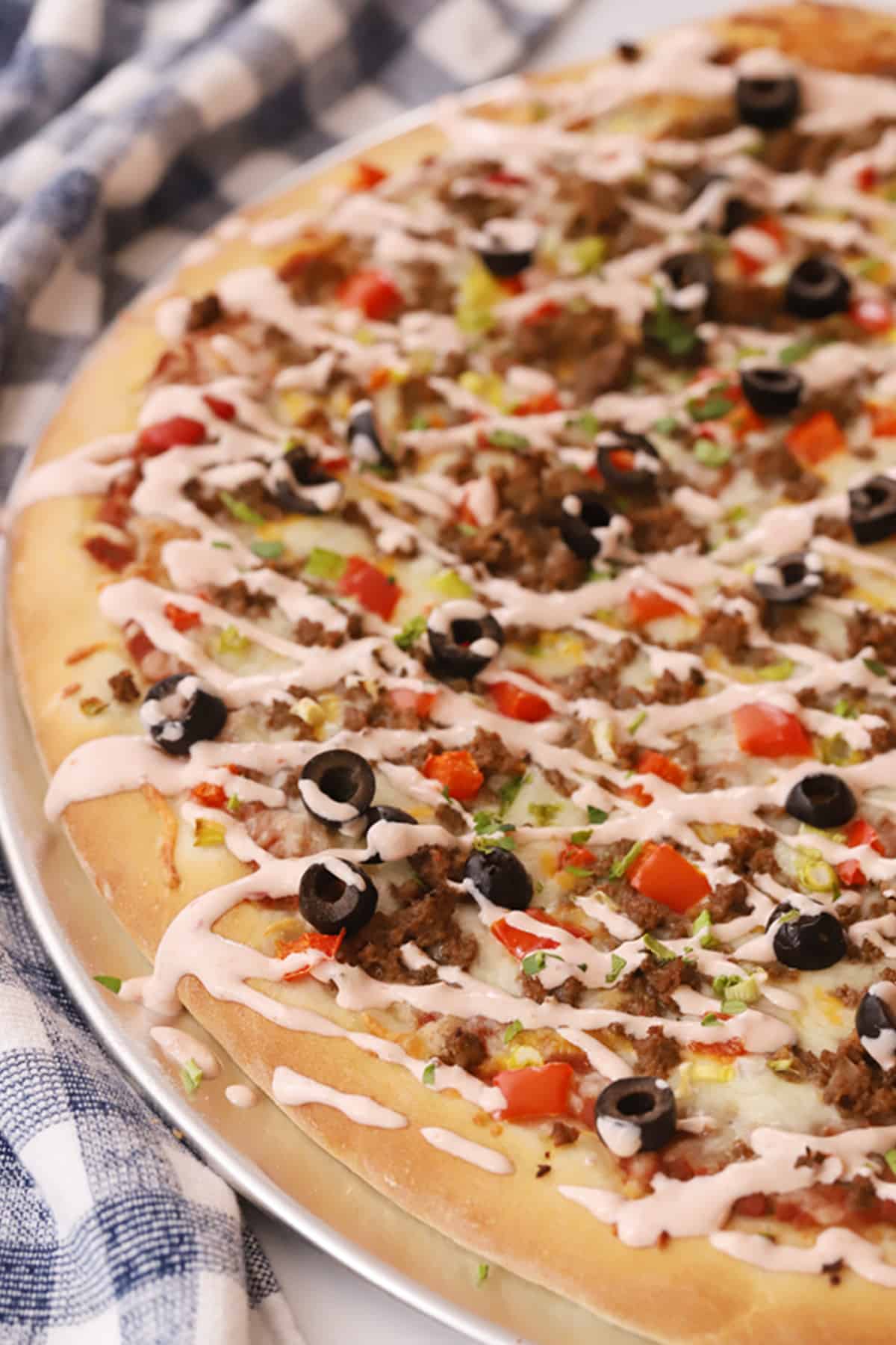 A vibrant homemade taco pizza, lavishly topped with seasoned ground beef, ripe olives, fresh tomatoes, crisp lettuce, and a delightful creamy Mexican drizzle, ready to be enjoyed.