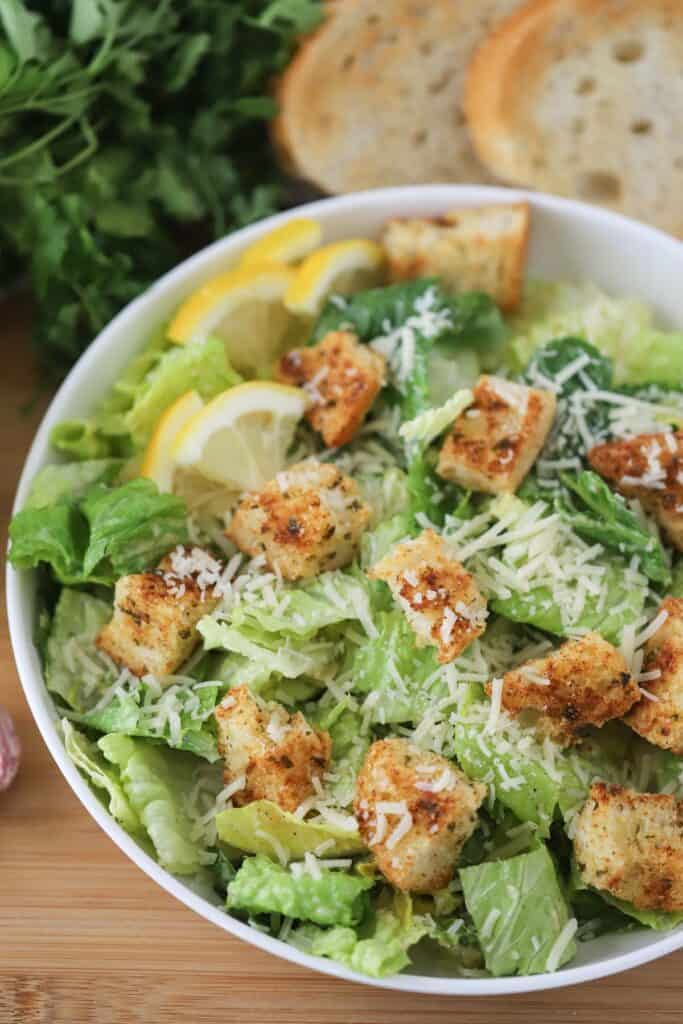 A white bowl full of the best caesar salad recipe, topped with homemade croutons, shaved parmesan cheese, and lemon wedges. 