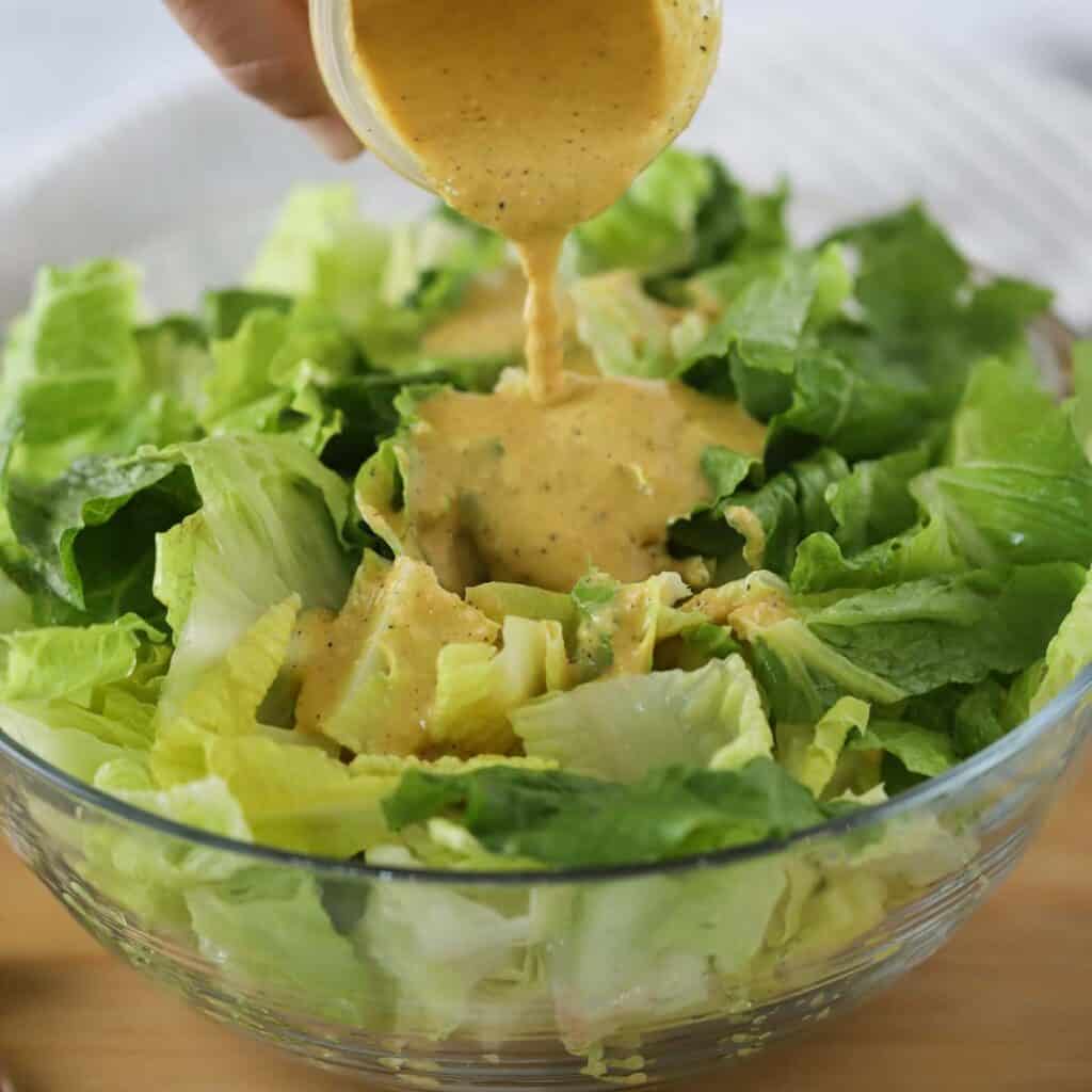 Creamy caesar dressing being poured over a large salad bowl full of romaine lettuce. This is the best Caesar salad recipe.