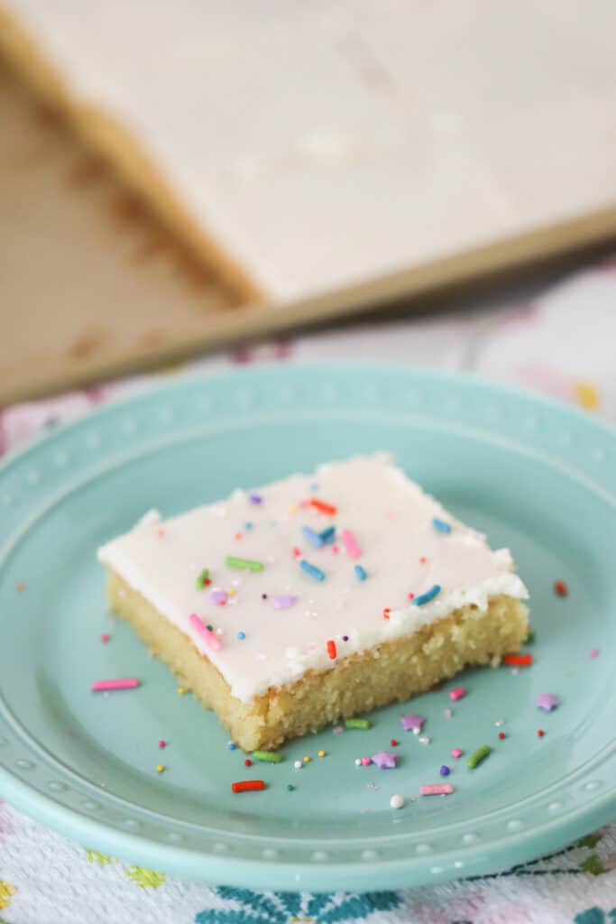 A slice of white Texas sheet cake on a plate, topped with colorful sprinkles and white frosting.
