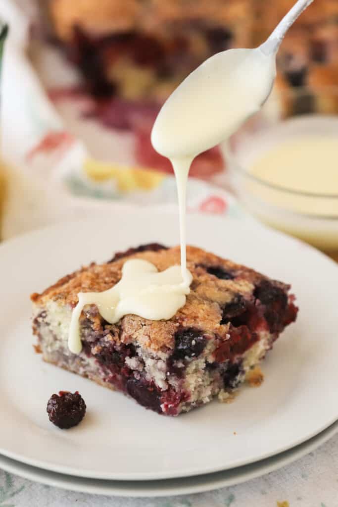 Mixed berry coffee cake on a white plate with vanilla glaze being poured over the top. The best breakfast cake recipe.