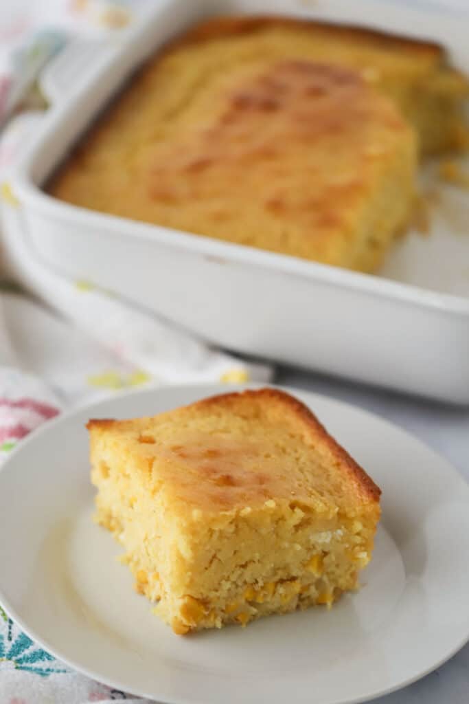 A slice of this creamed corn casserole recipe on a white serving plate with the full tray in the background.
