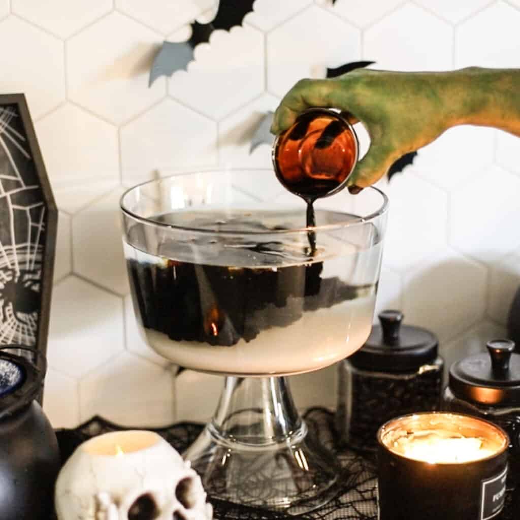 A spooky green hand adding root beer extract to a punch bowl surrounded by halloween decorations.