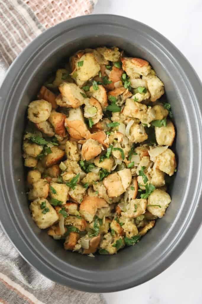 A slow cooker full of dry bread cubes and cooked onions and celery to make crockpot stuffing.