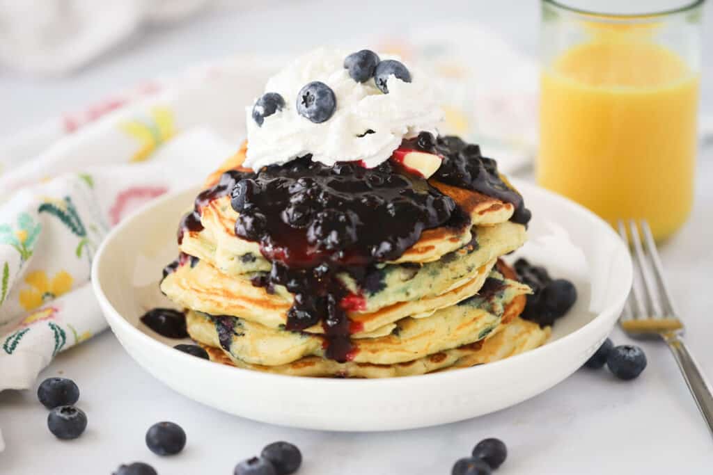 A breakfast plate filled with lemon blueberry pancakes topped with blueberry syrup and whipped cream.
