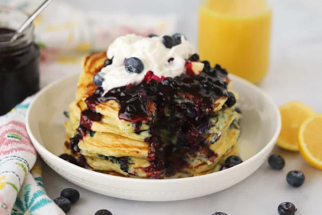 Homemade blueberry syrup poured over a stack of pancakes with whipped cream.